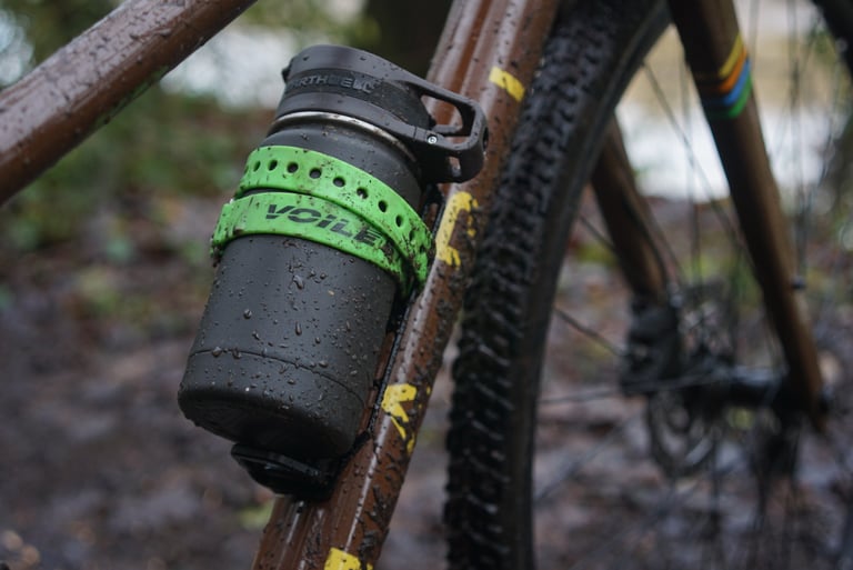 Tailfin Cargo Cage with Load Chip on a muddy brown steel frame, holding an oversized flask with a green Voile strap.