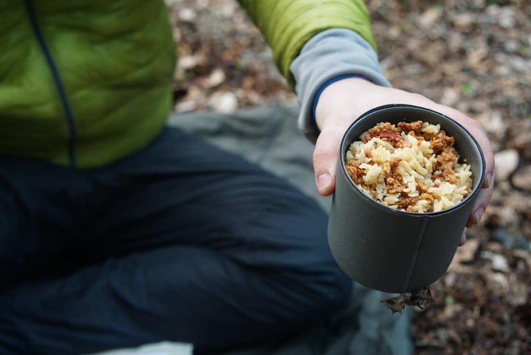 Rider in a green jacket holds out a mug of Firepot Vegan Orzo Bolognese at a woodland bikepacking camp.