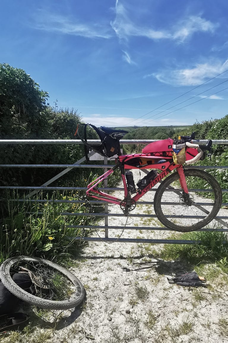 Pink Specialized Diverge with bikepacking bags hung on a metal field gate in Dorset, rear wheel off for a repair.