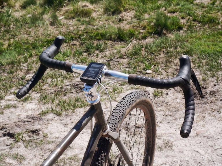 Wide Cowchipper Deluxe 52cm bars with a bike computer on a muddy bike, parked on a dry grassy track.