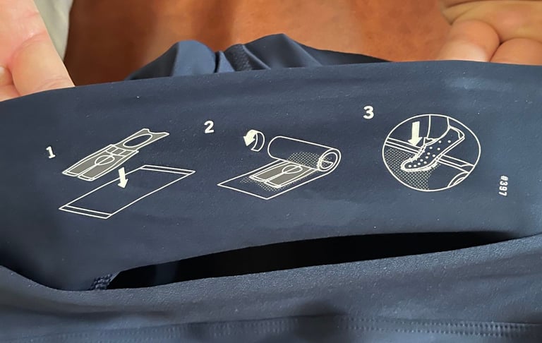 Three-step towel-rolling diagram inside the waistband of Rapha Core Cargo shorts, ending with a shoe pressing on the roll.