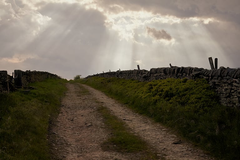Sunbeams break through cloud over a stony gravel track between dry stone walls in the Peak District.
