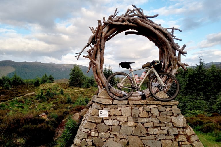 Bike propped inside a driftwood ring sculpture on a stone plinth beside the Badger Divide, with forested hills behind.