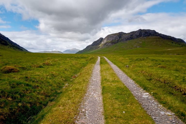 Double-track gravel path through a green glen below rocky hills, on the Badger Divide route down to Corrour.