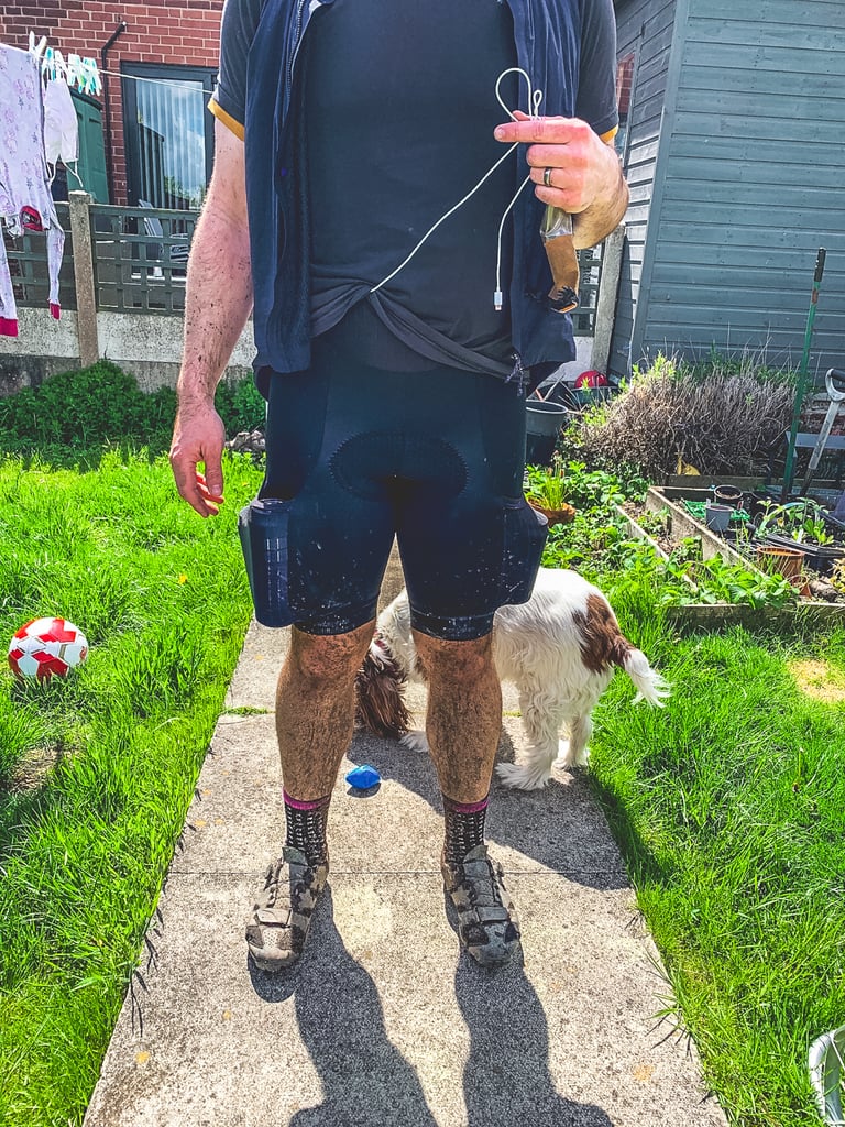 Muddy rider in Lusso Adventure bib shorts and cycling shoes stands on a garden path with a dog behind.