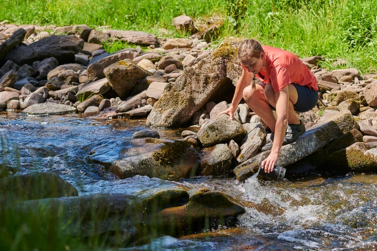 Gail Brown crouches on riverside rocks to fill a bottle from a clear, fast-flowing stream.