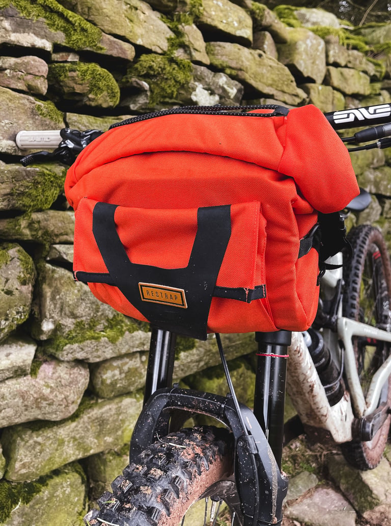 Orange Restrap Bar Pack on the bars of a muddy full-suspension mountain bike, against a mossy stone wall.