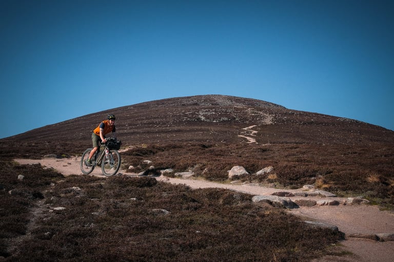 Bikepacker in olive shorts rides a loaded bike along a stony path below a bare hill on a Scottish bikepacking trip.