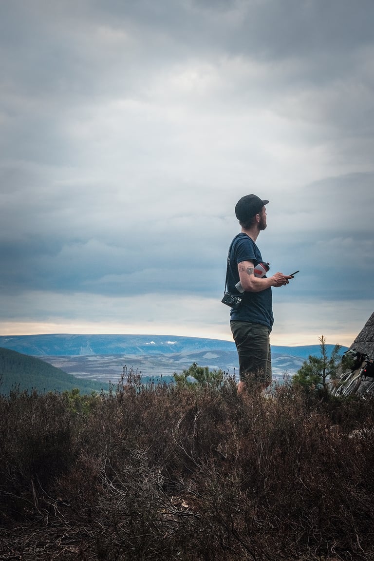 Rider in Vulpine shorts and a cap stands on heather moorland looking across Scottish hills, camera over shoulder.