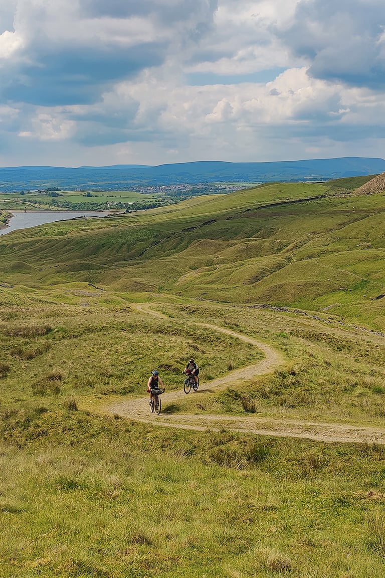 Two bikepackers ride a winding gravel track through green moorland above a reservoir during the bib short test.
