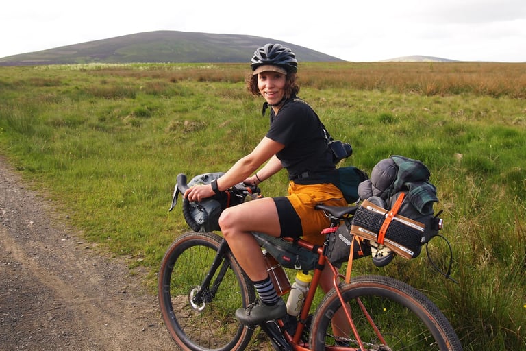 Smiling rider in orange shorts over the Pearl Izumi bibs, on a loaded bikepacking bike beside a moorland gravel track.