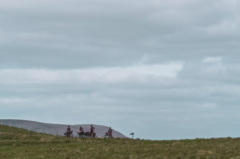 Four Pennine Rally riders with bikes stop on a distant ridge under a wide grey sky.