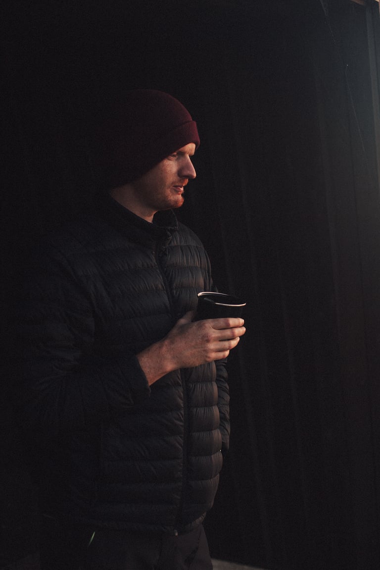 Camper in a beanie and down jacket cradles a mug in near darkness, early on the morning after the bivy.