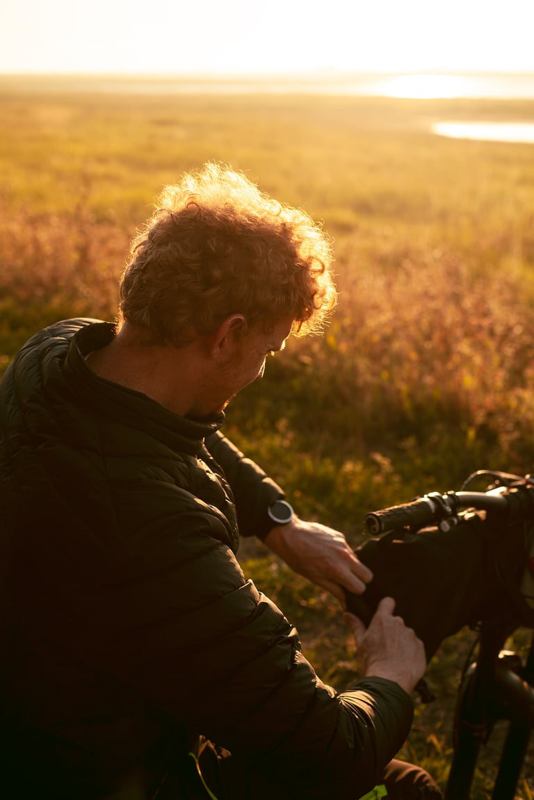 Rider crouched beside a handlebar bag in warm evening light, with marshland stretching away behind.