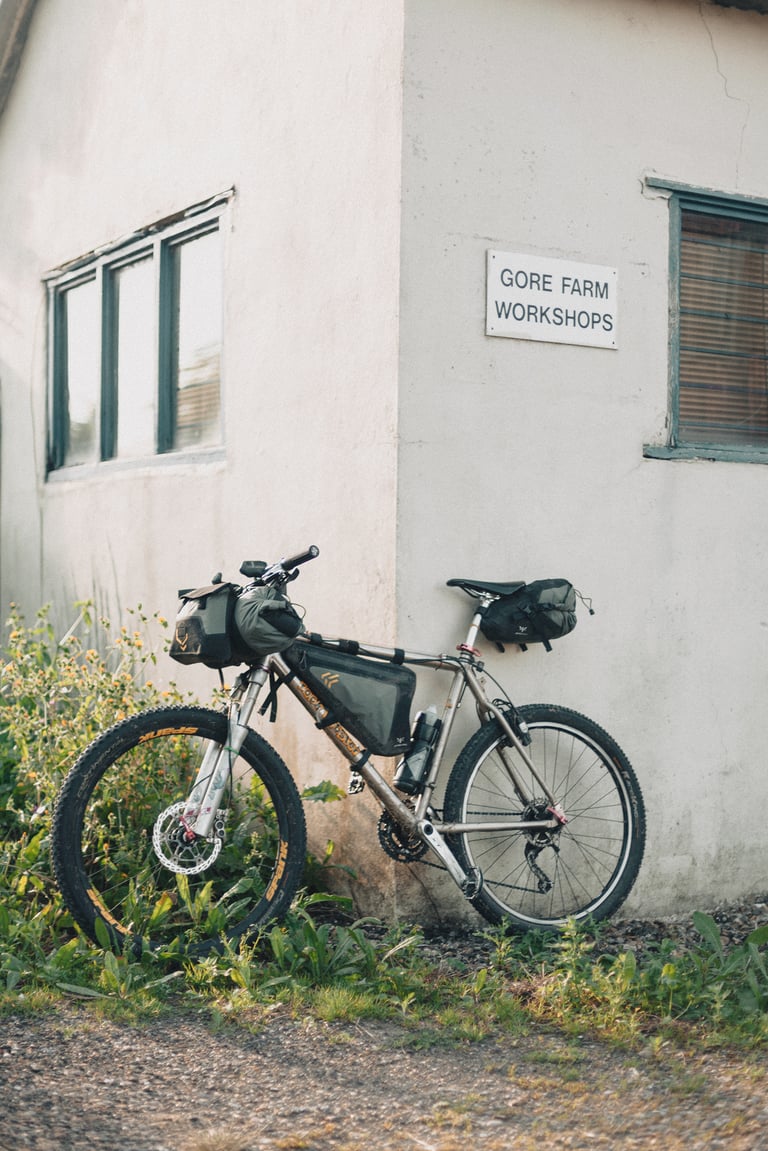 Mountain bike loaded with frame, handlebar and saddle bags leaning against a wall by a Gore Farm Workshops sign.