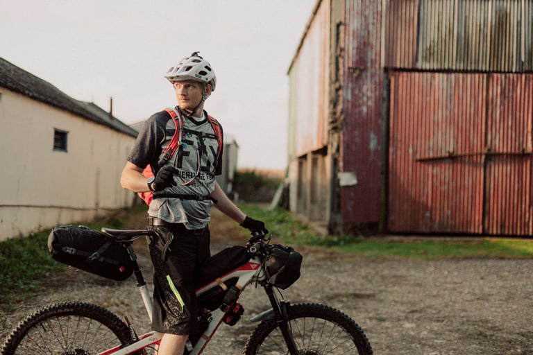 Rider in a helmet and hydration pack pauses beside a loaded mountain bike outside farm buildings at dusk.