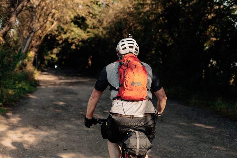Rider in a white helmet and orange hydration pack heads along a shaded gravel track on the way to a local overnighter.
