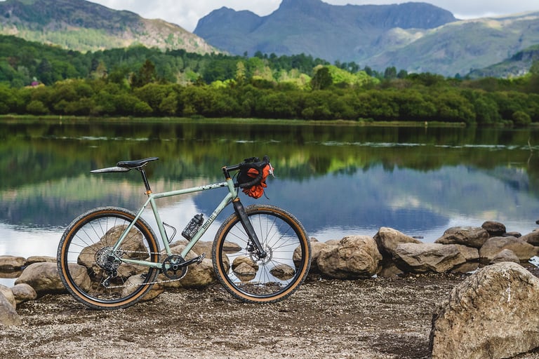 Pale green gravel bike with an Ashdown Bags bar bag and orange jacket, parked on a stony lakeshore below fells.