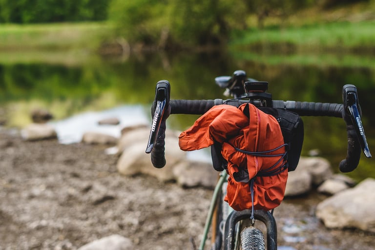 Orange rain jacket stuffed under the elastic cord of an Ashdown Bags bar bag on a gravel bike by the water.