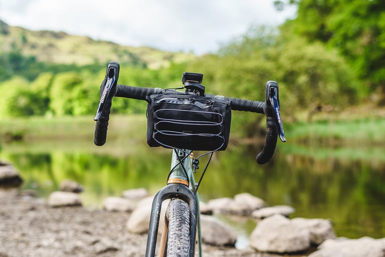Head-on view of the Ashdown Bags canvas bar bag between the drops of a gravel bike, with wooded hills and a lake behind.