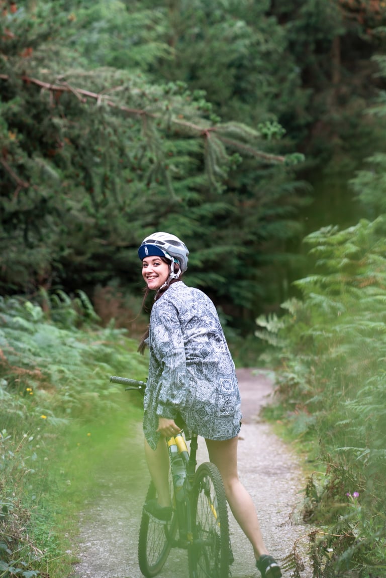 Immy looks back smiling, straddling her bike on a gravel track between bracken and pine trees.