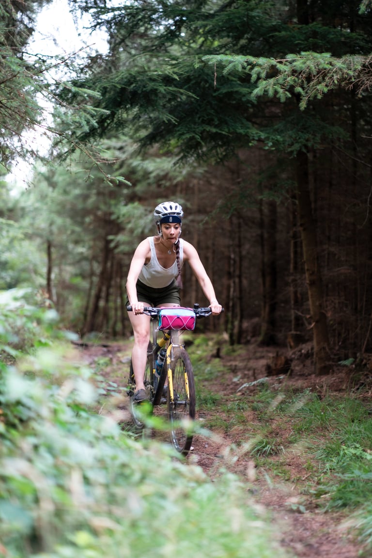 Immy rides a yellow mountain bike with a pink bar bag along a pine forest trail.