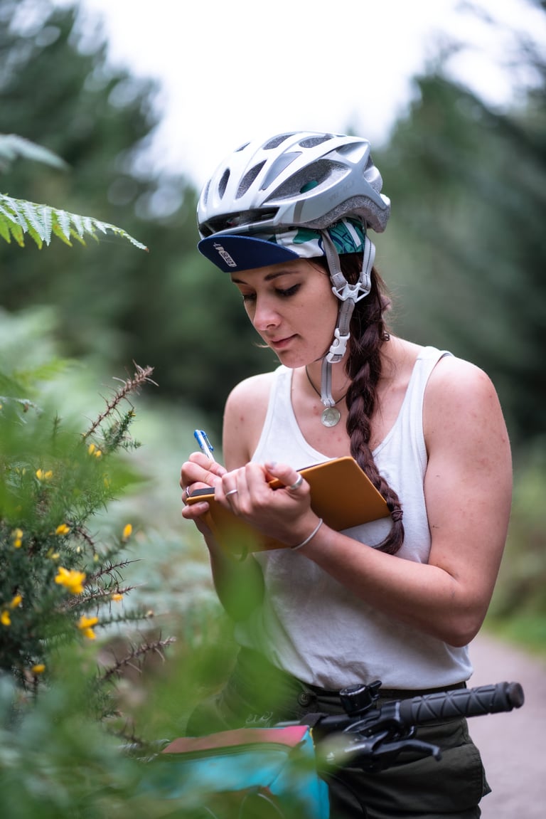Rider in a helmet pauses to write in an orange notebook beside flowering gorse, handlebar bag in front.