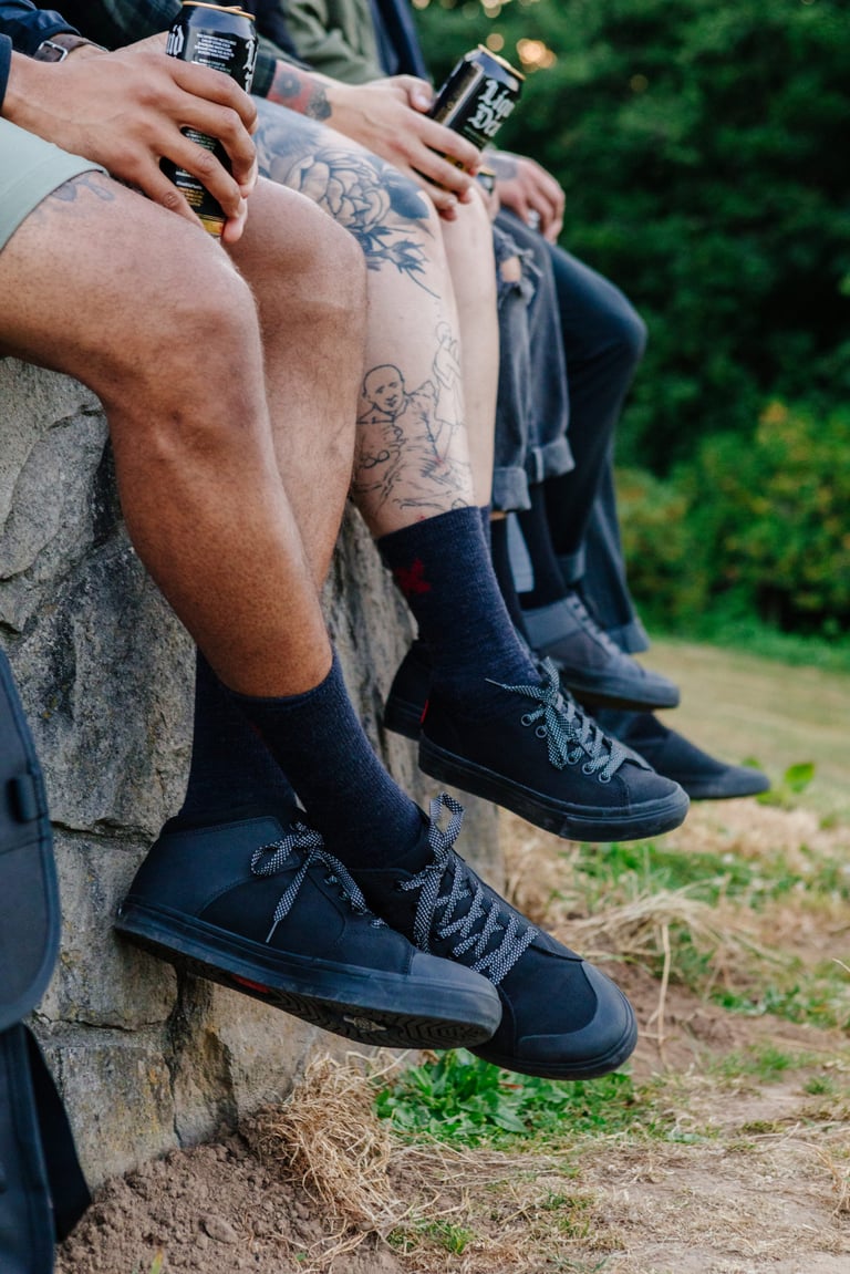 Three people sit on a stone wall with cans of beer, wearing black Chrome trainers from the Panaracer Pedal Series.