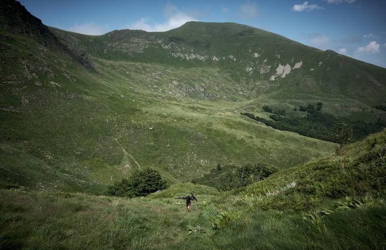 Lone rider shoulders a bike up a grassy mountain valley below rocky peaks.