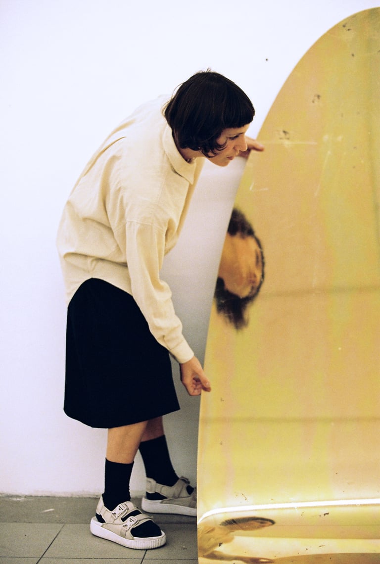 Quinda Verheul leans in to look at her reflection in a curved, gold-toned mirror sculpture against a white wall.