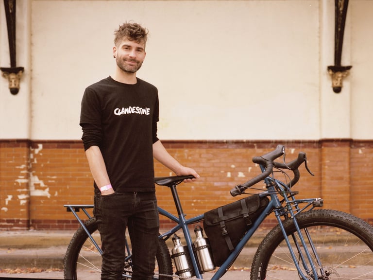 Pi Manson of Clandestine stands beside his blue Long Tail adventure bike with frame bag and steel bottles at Bespoked 2021.