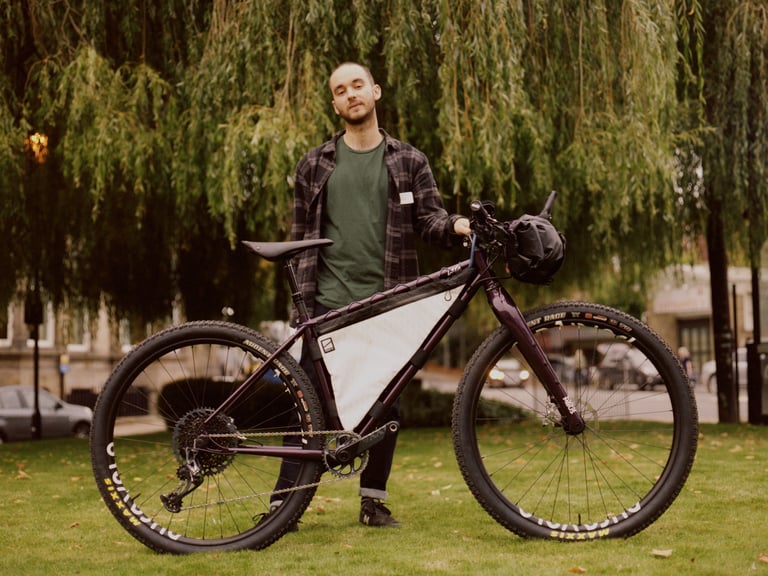 Chris Lord of Lord Cycles stands behind the Nomad, a purple rigid endurance mountain bike with a frame bag, at Bespoked.