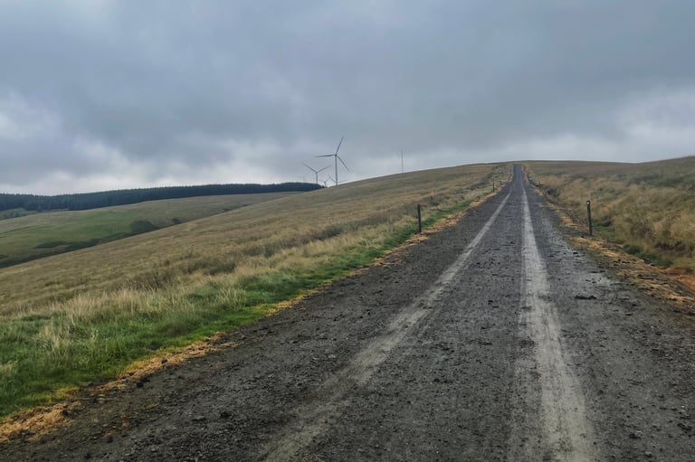 Wide gravel track climbs a grassy hillside towards wind turbines under grey cloud on the Second City Divide.