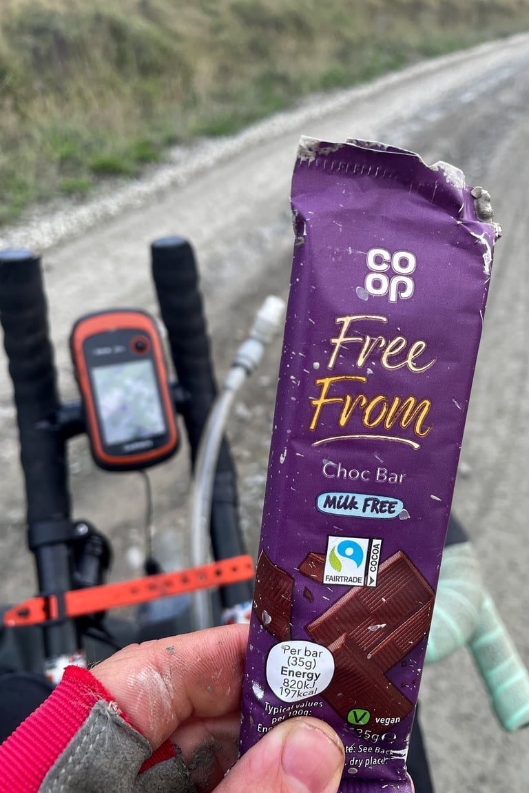 Hand holds a Co-op Free From vegan chocolate bar over bikepacking handlebars with a GPS unit on a gravel track.