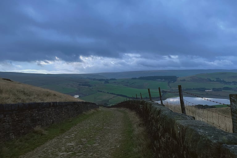 Stone-walled bridleway drops towards moorland reservoirs under heavy cloud on the Second City Divide in the Pennines.