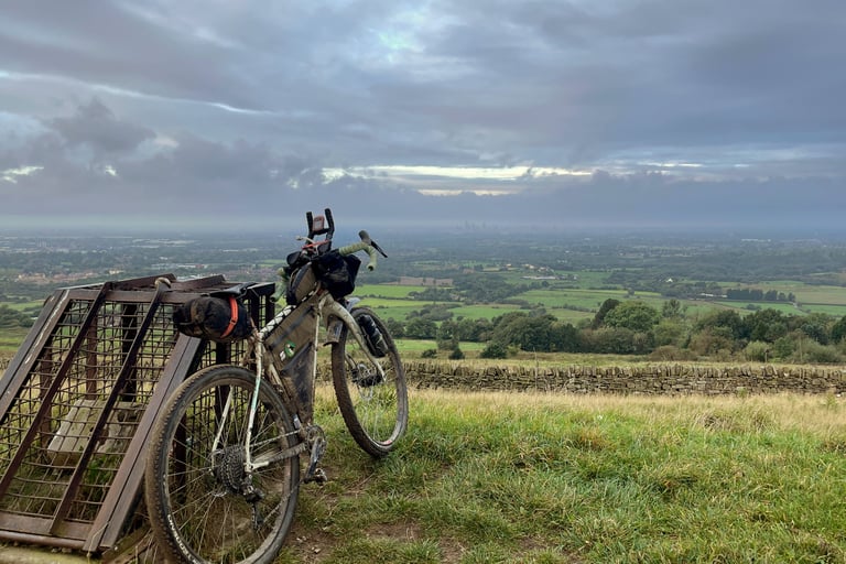 Loaded bikepacking bike leans on a rusty hilltop gate with the hazy Manchester skyline far across the plain.