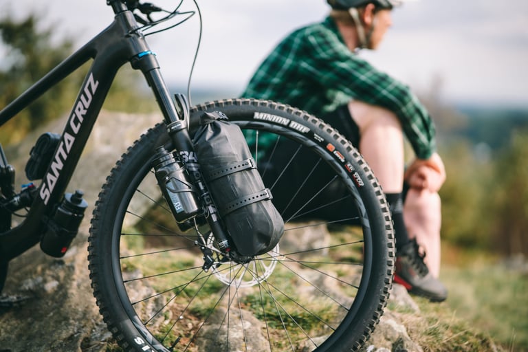 Tailfin fork mounts carrying a drybag and a bottle on one leg of a Santa Cruz mountain bike, rider seated behind.