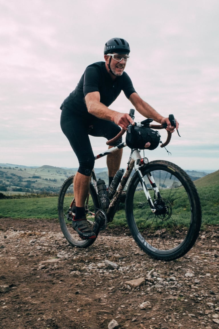 Smiling rider climbs a stony track on a gravel bike with a handlebar bag, hills behind, on the Gravel Bonanza.