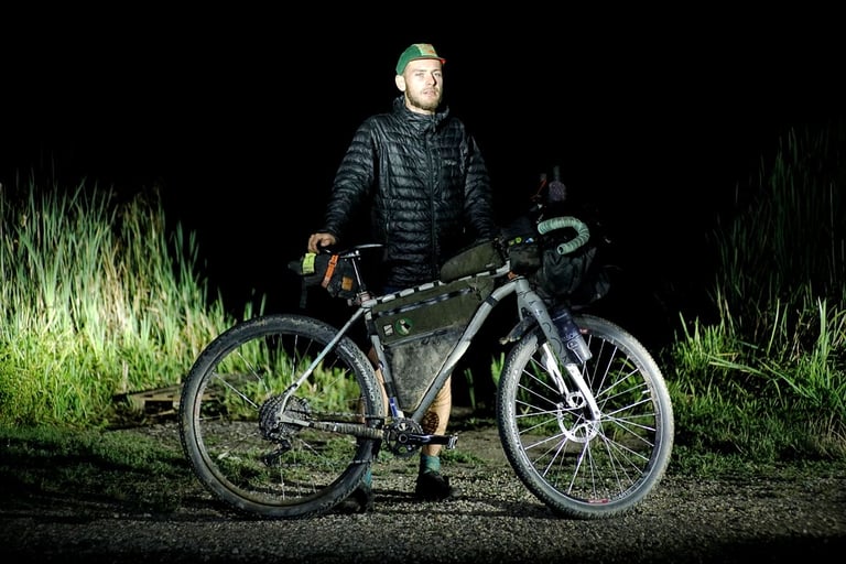 Marcus Nicolson stands at night beside his loaded bikepacking bike on gravel at the finish of the Hungarian Divide.