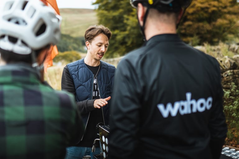 Man in a quilted gilet talks to two riders, one in a Wahoo jacket, at the Gravel Bonanza feed stop in the Goyt Valley.