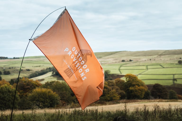 Orange Outdoor Provisions flag blowing in the wind above Peak District fields and hills at the Gravel Bonanza feed stop.