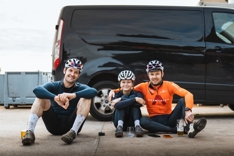 Three muddy-legged riders in helmets sit on the ground by a black van with pints at the Service Course Gravel Bonanza finish.