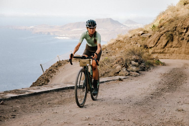 Rider on a green Orbea Terra with a handlebar bag climbs a dirt road along a rocky hillside above the sea.