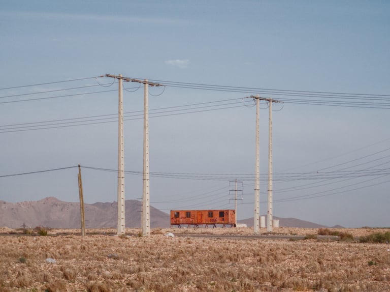 Concrete pylons and power lines cross arid scrub outside Marrakech, an orange container and bare hills behind.