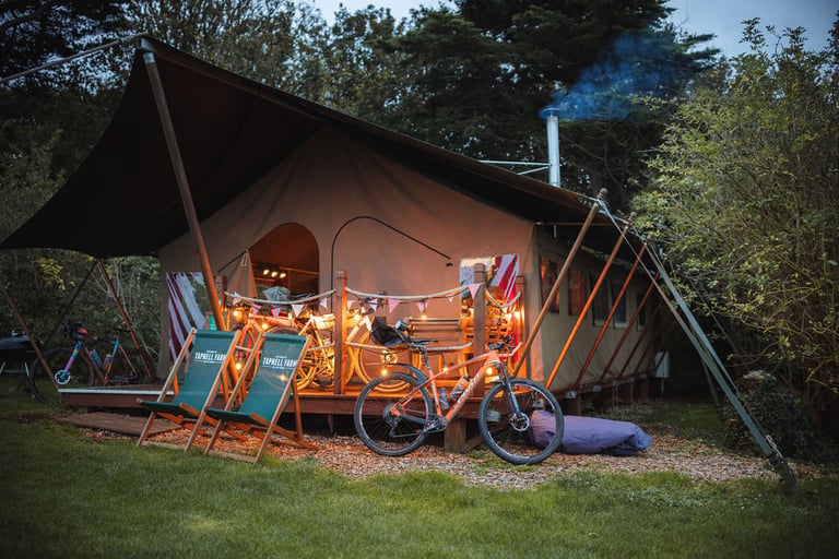 Lit-up safari tent at Tapnell Farm at dusk with deckchairs and bikes parked on the veranda and lawn.
