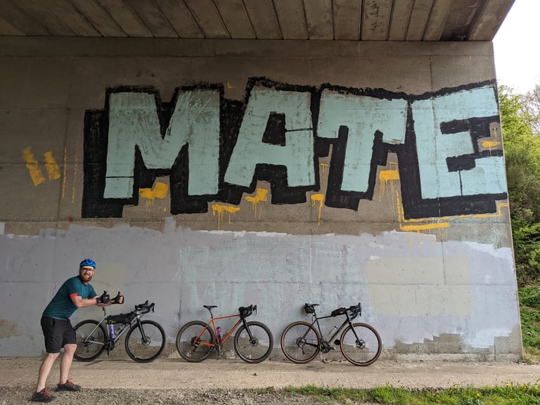 Rider gives two thumbs up beside three Fairlight Secans in silver, orange and black under a bridge with MATE graffiti.