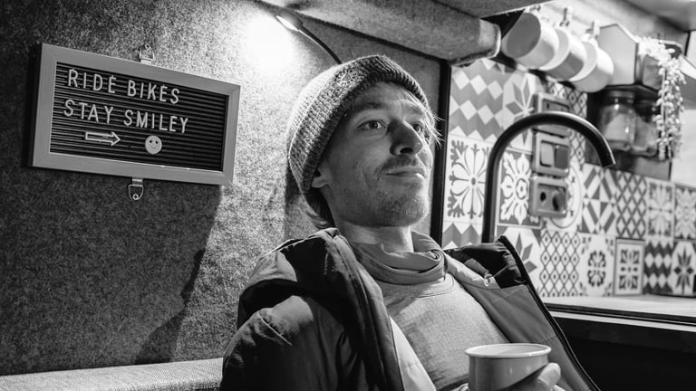 Black and white portrait of a smiling man in a beanie holding a cup, beside a Ride Bikes Stay Smiley sign.