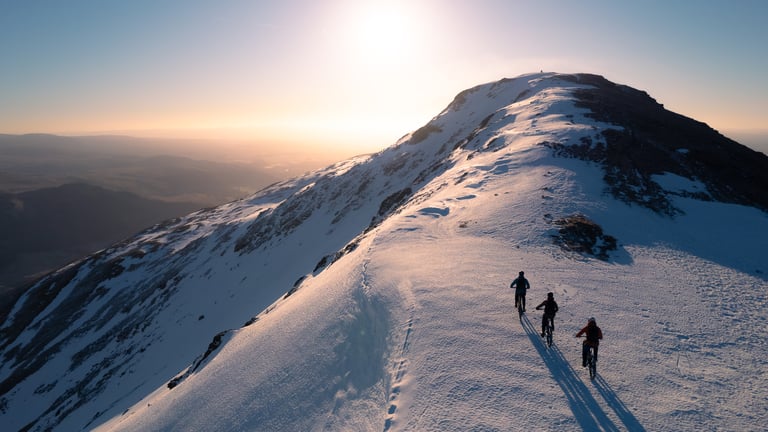 Three riders climb a snow-covered mountain ridge towards the summit in low sun, long shadows behind them.