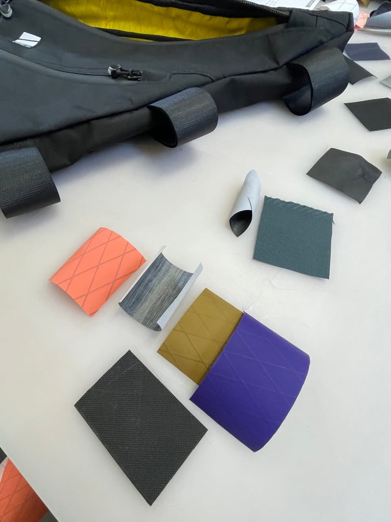 Fabric swatches in orange, purple, mustard and grey beside a black frame bag, for choosing the bag's materials.