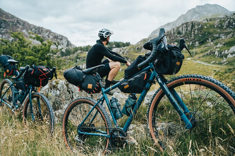 Rider sits on a rock between two Ribble Gravel 725s loaded with bikepacking bags, rocky hills behind.